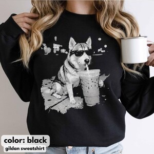 May include: Black Gildan sweatshirt featuring a grayscale graphic of a husky wearing sunglasses and drinking from a clear iced coffee cup. The text "color: black" and "gildan sweatshirt" are at the bottom.