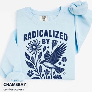 May include: Light blue sweatshirt with the words "RADICALIZED BY" above a dark blue floral and bird graphic. The sweatshirt has a crew neck and long sleeves. The word "CHAMBRAY" is printed at the bottom.