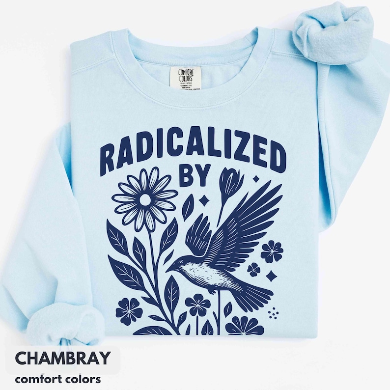 May include: Light blue sweatshirt with the words "RADICALIZED BY" above a dark blue floral and bird graphic. The sweatshirt has a crew neck and long sleeves. The word "CHAMBRAY" is printed at the bottom.