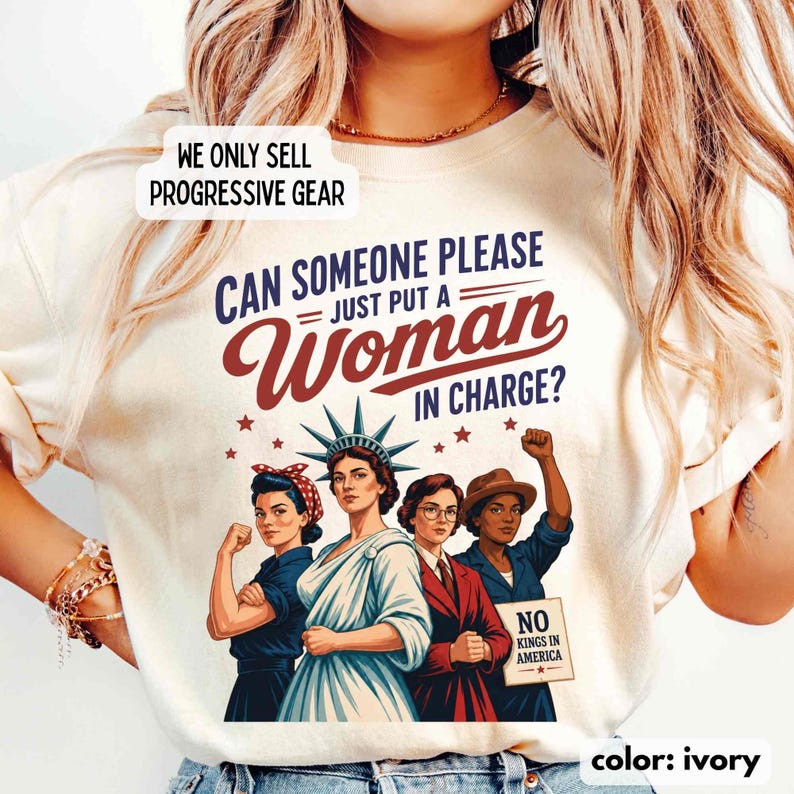 May include: Ivory-colored t-shirt with the text "CAN SOMEONE PLEASE JUST PUT A WOMAN IN CHARGE?" and an illustration of four women. The shirt also features the text "NO KINGS IN AMERICA" and "WE ONLY SELL PROGRESSIVE GEAR".