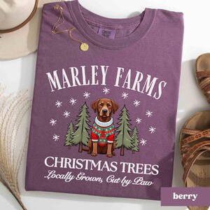 K&ouml;nnte beinhalten: Lilafarbenes T-Shirt mit einer wei&szlig;en Grafik eines braunen Labrador Retrievers, der einen rot-gr&uuml;nen Weihnachtspullover tr&auml;gt. Die Grafik enth&auml;lt auch Kiefern und Schneeflocken. Der Text auf dem Shirt lautet "Marley Farms Christmas Trees Locally Grown, Cut by Paw."