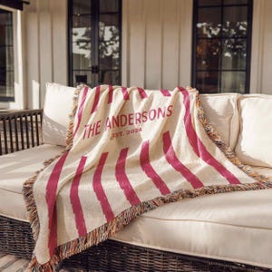 May include: Cream-colored throw blanket with vertical magenta stripes and fringed edges. The text "THE ANDERSONS EST. 2024" is printed in magenta. The blanket is draped over a white sofa on a patio.
