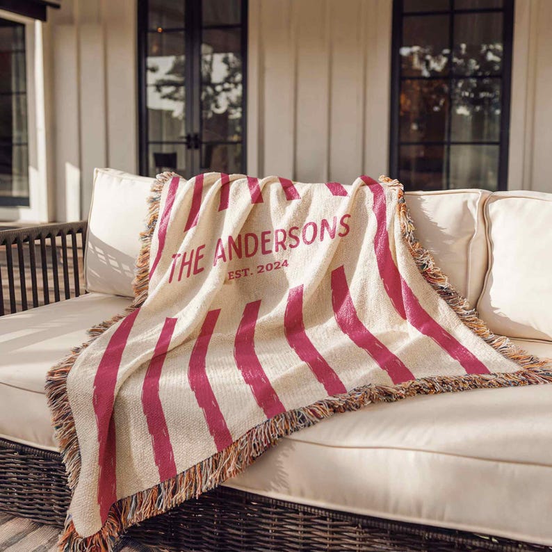 May include: Cream-colored throw blanket with vertical magenta stripes and fringed edges. The text "THE ANDERSONS EST. 2024" is printed in magenta. The blanket is draped over a white sofa on a patio.