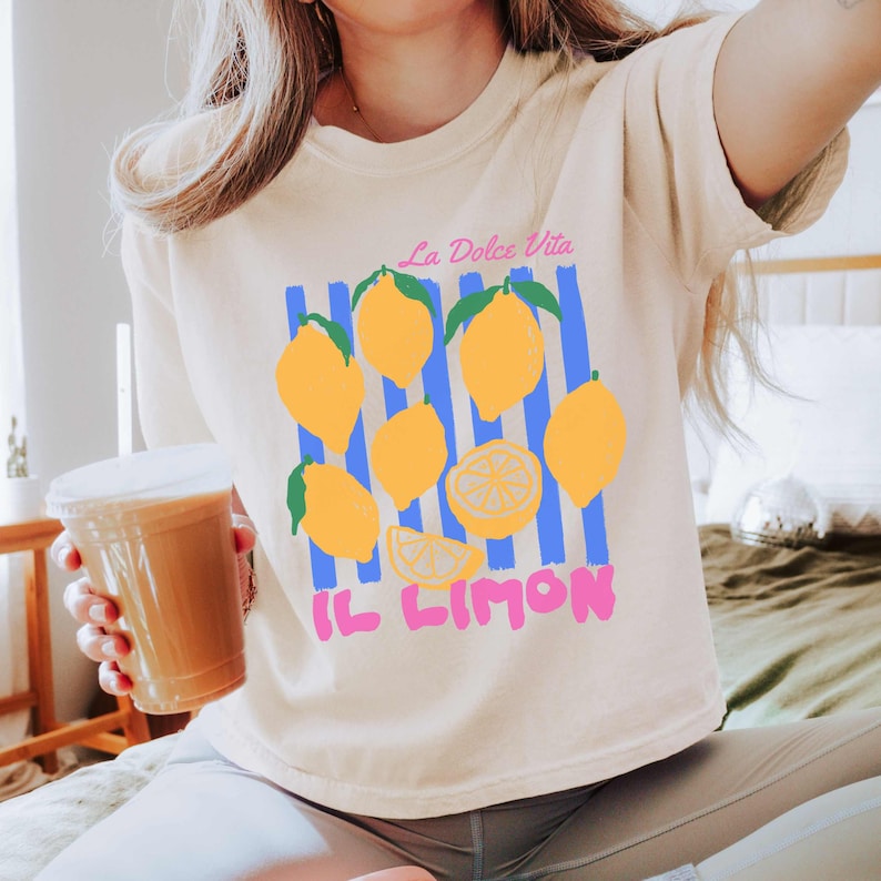 May include: A cream-colored t-shirt featuring a lemon-themed graphic. The design includes yellow lemons, blue vertical stripes, and the Italian text "La Dolce Vita" and "Il Limon" in pink. The shirt is a casual, everyday wear item.