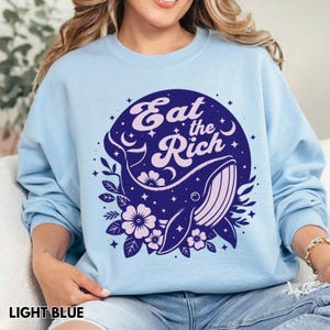 May include: Light blue sweatshirt featuring a whale graphic with stars and the text "Eat the Rich" in a circle.