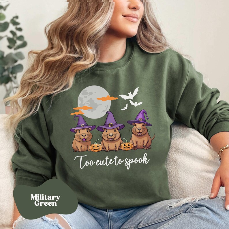 Op de afbeelding: Een groen sweatshirt met een afbeelding van drie cavia's die heksenhoeden dragen en pompoenen vasthouden. De tekst "Too cute to spook" staat onder de cavia's.