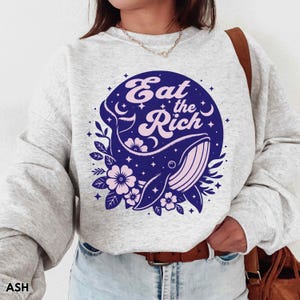May include: A grey sweatshirt featuring a blue and pink graphic of a whale with stars and flowers. The graphic reads "Eat the Rich" in pink lettering.