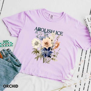 May include: Orchid-colored t-shirt with the text "ABOLISH ICE" above a floral bouquet in shades of white, blue, and peach. The shirt is paired with denim jeans and a green and white checkered hair clip.