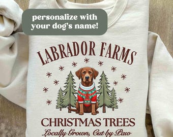 Op de afbeelding: Een wit sweatshirt met een bruine Labrador Retriever die een rood-groene kersttrui draagt voor een kerstboom. De tekst "LABRADOR FARMS CHRISTMAS TREES Locally Grown, Cut by Paw" staat op het sweatshirt gedrukt.