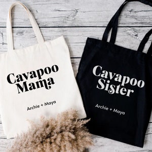 May include: Two tote bags, one white and one black, with the text "Cavapoo Mama" and "Cavapoo Sister" in black and white lettering, respectively. Both bags have the text "Archie + Maya" underneath the main text.