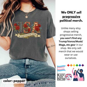 Op de afbeelding: Pepperkleurig T-shirt met het woord "USA" in een patriottisch ontwerp met rode rozen en blauwe accenten. De tekst "250 YEARS OF RESISTING FASCISM" staat op een banner onder het woord "USA". Het shirt maakt deel uit van een progressieve politieke merch collectie.