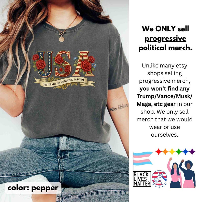 Op de afbeelding: Pepperkleurig T-shirt met het woord "USA" in een patriottisch ontwerp met rode rozen en blauwe accenten. De tekst "250 YEARS OF RESISTING FASCISM" staat op een banner onder het woord "USA". Het shirt maakt deel uit van een progressieve politieke merch collectie.