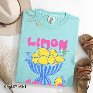 May include: A light blue t-shirt with the word "LIMON" in pink, and a graphic of lemons in a blue bowl. The text "la dolce vita" is below the word "LIMON". The shirt is labeled "CHALKY MINT" and is size XL-TO-80.