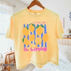 May include: A yellow t-shirt with a lemon graphic. The graphic includes yellow lemons, blue stripes, and the text "La Dolce Vita" and "Il Limon" in pink. The shirt is hanging on a wooden hanger.