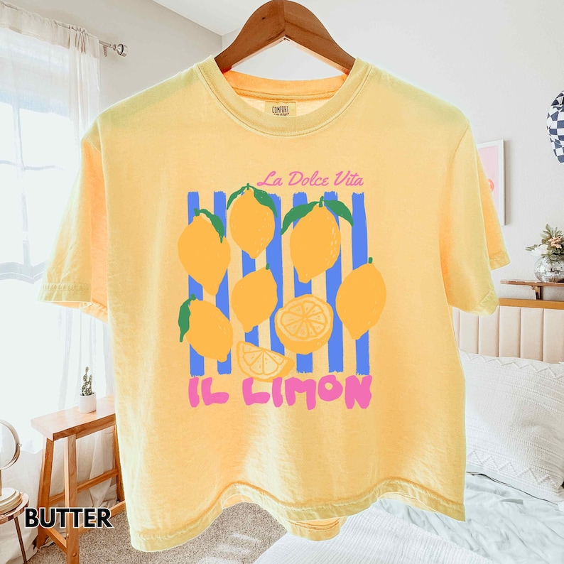 May include: A yellow t-shirt with a lemon graphic. The graphic includes yellow lemons, blue stripes, and the text "La Dolce Vita" and "Il Limon" in pink. The shirt is hanging on a wooden hanger.