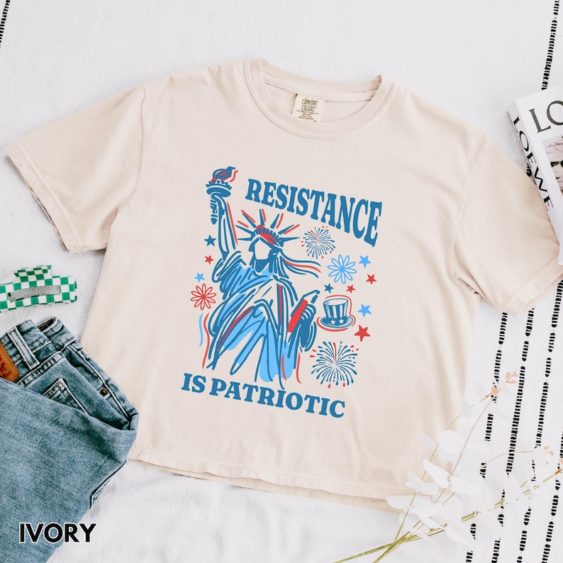 May include: Ivory-colored t-shirt with a graphic of the Statue of Liberty in red and blue, surrounded by stars and fireworks. The text "RESISTANCE IS PATRIOTIC" is printed above and below the image. The shirt is paired with blue jeans.