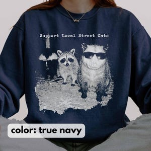 May include: True navy sweatshirt with a graphic of a raccoon and an opossum wearing sunglasses. The text "Support Local Street Cats" is printed above the graphic. The bottom of the sweatshirt has the text "color: true navy".
