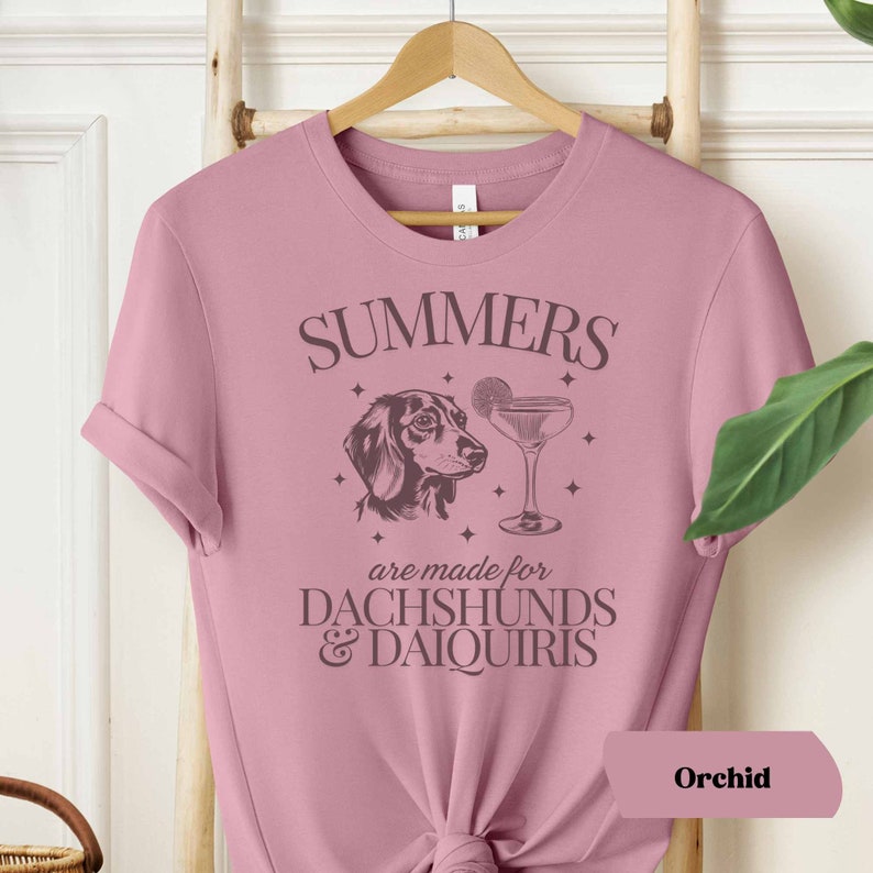 May include: A pink t-shirt with the text "SUMMERS are made for  DACHSHUNDS & DAIQUIRIS" in black. A black and white illustration of a dachshund and a cocktail glass are above the text. The text "Orchid" is in the bottom right corner.
