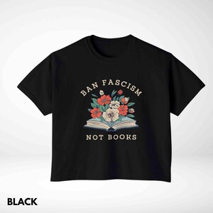 Op de afbeelding: Zwart T-shirt met de tekst "BAN FASCISM NOT BOOKS" boven een bloemenillustratie. Het ontwerp toont rode, witte en perzik kleurige bloemen die uit een open boek komen. Het woord "BLACK" staat onderaan.