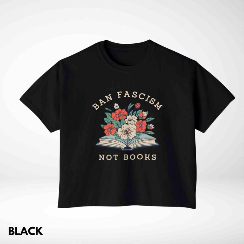Op de afbeelding: Zwart T-shirt met de tekst "BAN FASCISM NOT BOOKS" boven een bloemenillustratie. Het ontwerp toont rode, witte en perzik kleurige bloemen die uit een open boek komen. Het woord "BLACK" staat onderaan.