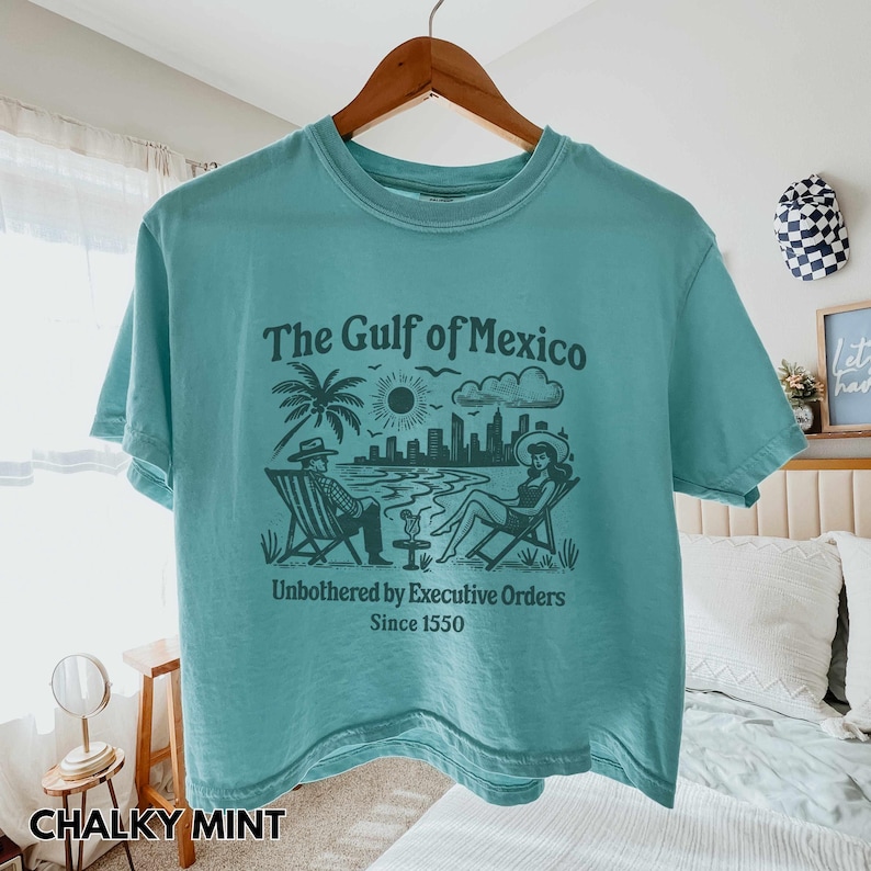 May include: A teal blue cropped t-shirt with a graphic print of a beach scene with the text "The Gulf of Mexico" and "Unbothered by Executive Orders Since 1550".