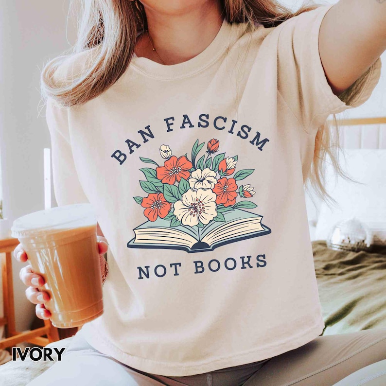 Op de afbeelding: Ivoor kleurig t-shirt met de tekst "BAN FASCISM NOT BOOKS" in marineblauw. Het shirt heeft een bloemenontwerp boven een open boek. De bloemen zijn rood, wit en oranje met groene bladeren. Het shirt is een casual stijl.