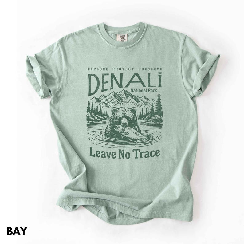 May include: A light green t-shirt with a brown bear holding a fish. The text "Explore Protect Preserve Denali National Park Leave No Trace" is printed on the shirt.