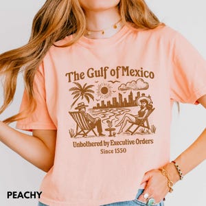 May include: Peach colored t-shirt with a brown graphic of a beach scene with the text "The Gulf of Mexico" and "Unbothered by Executive Orders Since 1550".