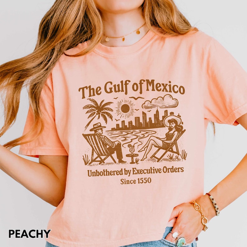 May include: Peach colored t-shirt with a brown graphic of a beach scene with the text "The Gulf of Mexico" and "Unbothered by Executive Orders Since 1550".