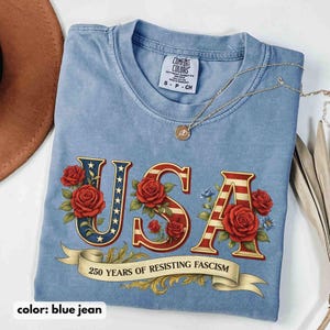 Op de afbeelding: Lichtblauwe spijkerbroekkleurige t-shirt met het woord "USA" in een patriottisch ontwerp, met rode rozen en een banner met de tekst "250 YEARS OF RESISTING FASCISM". Een gouden ketting ligt over het shirt.
