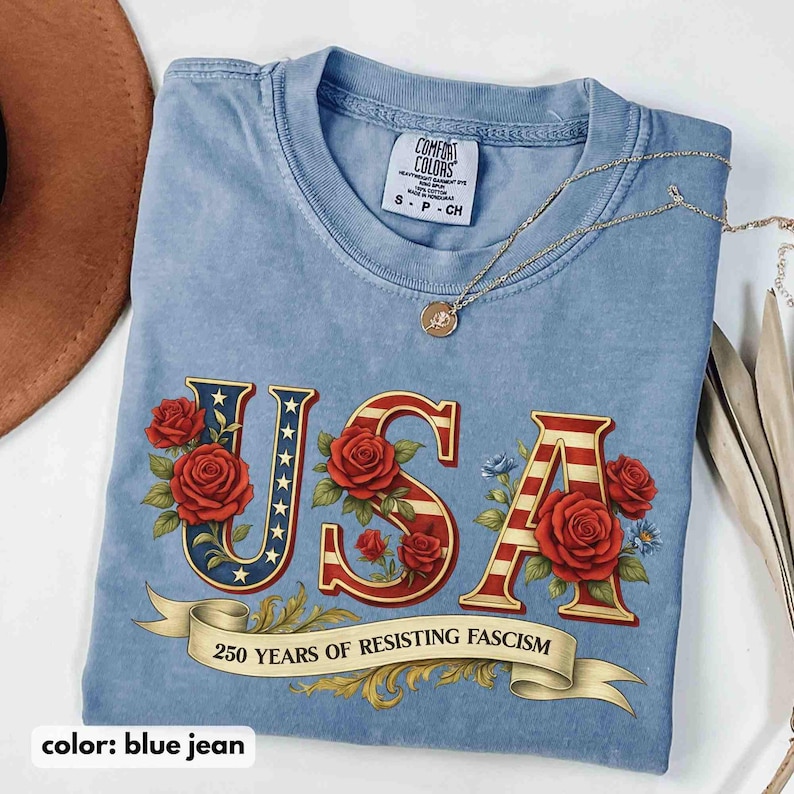 Op de afbeelding: Lichtblauwe spijkerbroekkleurige t-shirt met het woord "USA" in een patriottisch ontwerp, met rode rozen en een banner met de tekst "250 YEARS OF RESISTING FASCISM". Een gouden ketting ligt over het shirt.