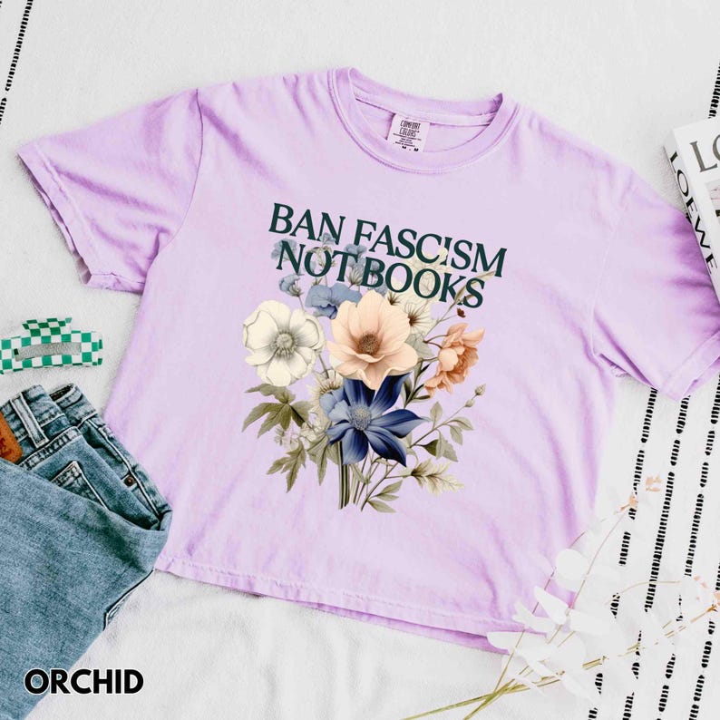 Puede incluir: Camiseta color orqu&iacute;dea con el texto "BAN FASCISM NOT BOOKS" en verde oscuro. Un ramo floral en tonos blancos, azules y melocot&oacute;n est&aacute; impreso debajo. La camiseta se combina con vaqueros y una pinza para el pelo verde y blanca. La palabra "ORCHID" est&aacute; impresa.