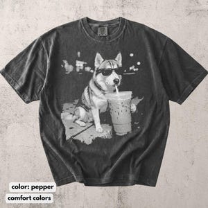May include: Pepper-colored t-shirt featuring a graphic of a husky wearing sunglasses and drinking from a large iced coffee cup. The dog is sitting on a sidewalk. The shirt has a vintage, washed look.