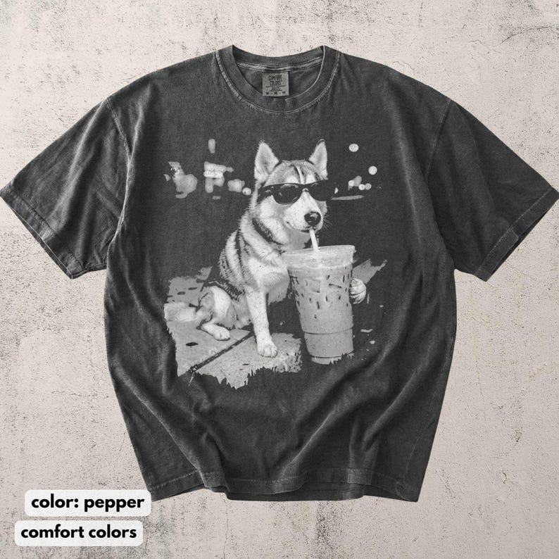 May include: Pepper-colored t-shirt featuring a graphic of a husky wearing sunglasses and drinking from a large iced coffee cup. The dog is sitting on a sidewalk. The shirt has a vintage, washed look.