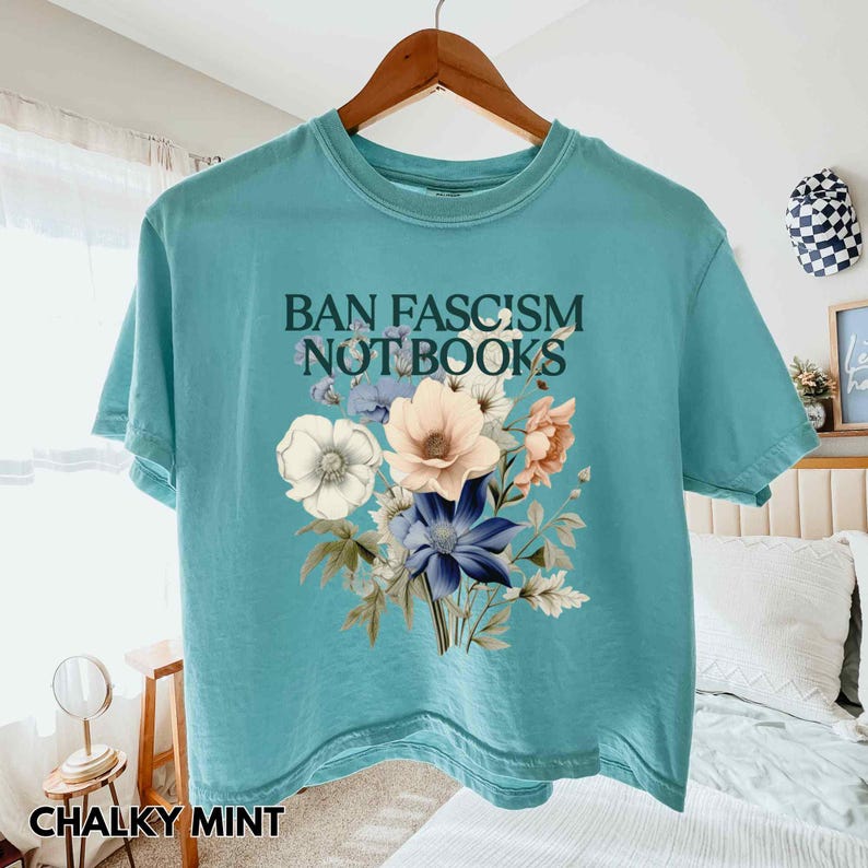 Puede incluir: Una camiseta turquesa con el texto "BAN FASCISM NOT BOOKS" sobre un ramo floral. El ramo presenta flores blancas, melocot&oacute;n y azules con hojas verdes. La camiseta est&aacute; colgada en una percha de madera.