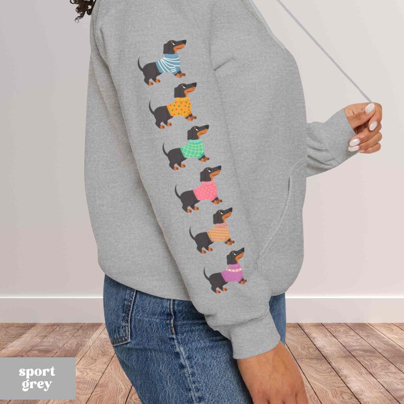 May include: A gray hooded sweatshirt with a row of black dachshund dogs wearing colorful sweaters printed on the sleeve. The text "sport grey" is printed on the bottom left corner of the image.