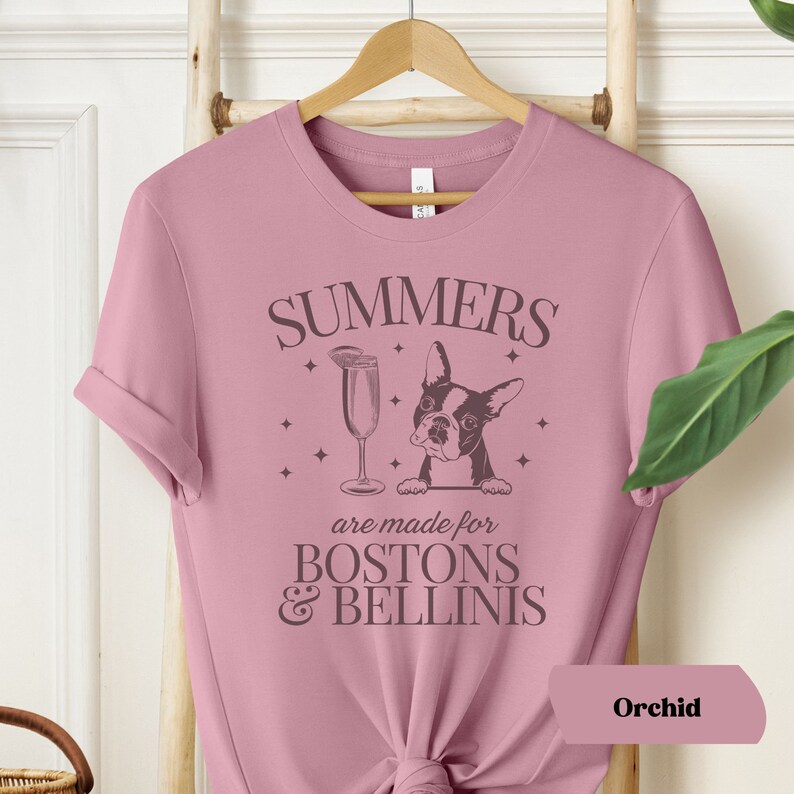 Puede incluir: Una camiseta rosa con el texto "SUMMERS are made for BOSTON & BELLINIS" y una ilustraci&oacute;n en blanco y negro de un Boston Terrier asom&aacute;ndose por encima del texto. El texto est&aacute; en una fuente marr&oacute;n.