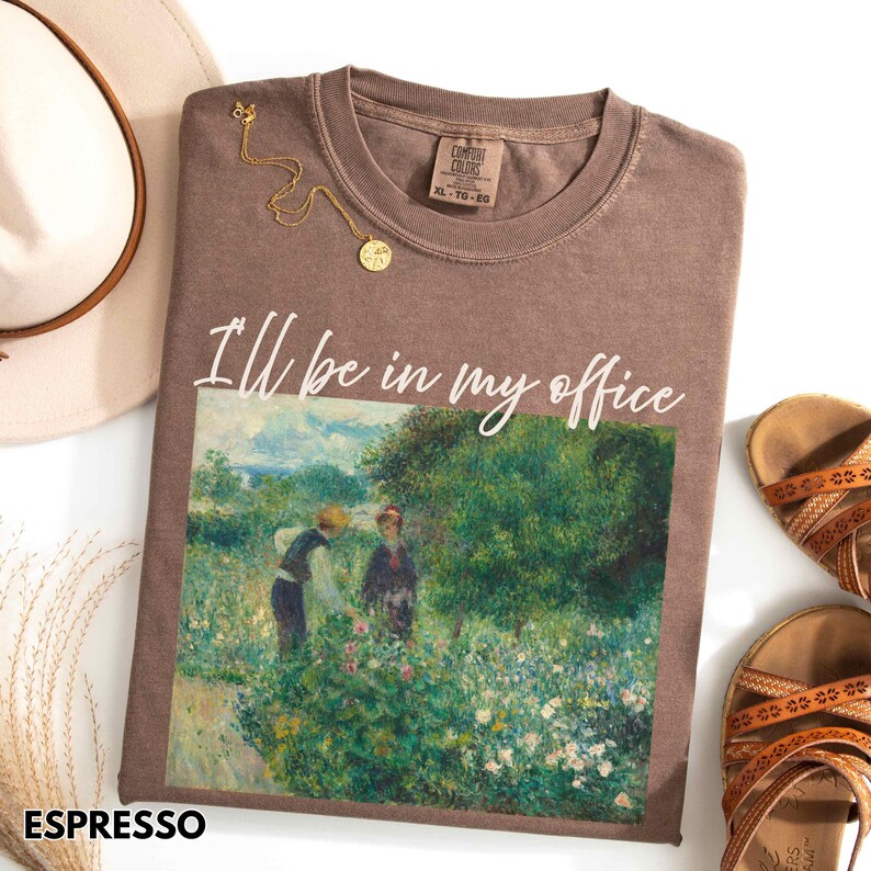 May include: Espresso-coloured t-shirt with the phrase "I'll be in my office" above a painting of people in a garden. The shirt has a crew neck and short sleeves. A gold necklace and sandals are next to the shirt.