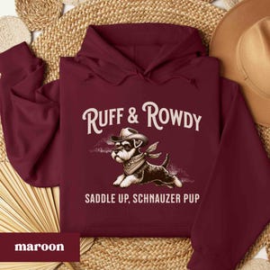 May include: A maroon hoodie with a graphic of a miniature schnauzer wearing a cowboy hat and bandana. The text "Ruff & Rowdy" and "Saddle Up, Schnauzer Pup" is printed on the hoodie.