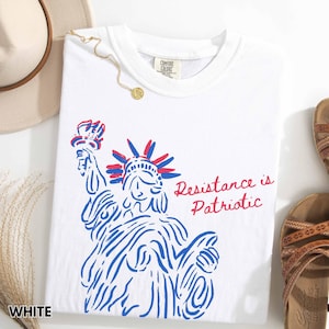 Pu&ograve; includere: T-shirt bianca con grafica blu e rossa della Statua della Libert&agrave; e la scritta "Resistance is Patriotic". La maglietta ha un girocollo e un'etichetta "Comfort Colors".