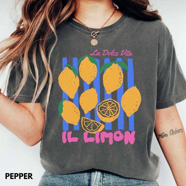 May include: A dark gray t-shirt with a colorful lemon graphic. The design includes yellow lemons, blue vertical stripes, and the text "La Dolce Vita" and "IL LIMON" in pink. The shirt is made of a soft material.