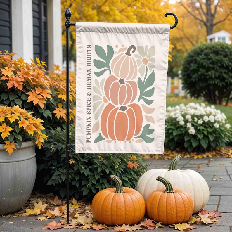 May include: A cream-colored garden flag with a stacked pumpkin design in shades of orange, peach, and green, with the text "PUMPKIN SPICE & HUMAN RIGHTS." Three pumpkins sit in front of the flag.
