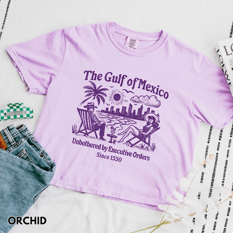 May include: A purple t-shirt with a graphic print of a beach scene with two people sitting in chairs under a palm tree. The text on the shirt reads "The Gulf of Mexico Unbothered by Executive Orders Since 1550".