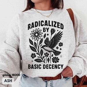 May include: Ash-colored sweatshirt with a black graphic of a bird, flowers, and the text "RADICALIZED BY BASIC DECENCY." The sweatshirt is a Gildan 18000. The design is centered on the front of the shirt.
