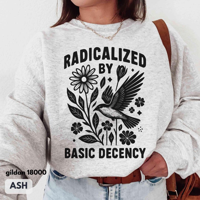 May include: Ash-colored sweatshirt with a black graphic of a bird, flowers, and the text "RADICALIZED BY BASIC DECENCY." The sweatshirt is a Gildan 18000. The design is centered on the front of the shirt.