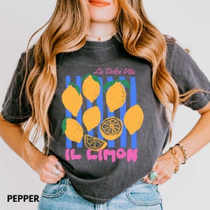 May include: Pepper-colored t-shirt with a lemon graphic. The design includes yellow lemons, blue stripes, and the text "La Dolce Vita" and "Il Limon" in pink. The shirt is a casual, vintage-style garment.