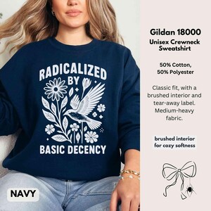May include: Navy blue crewneck sweatshirt with the text "RADICALIZED BY BASIC DECENCY" in white. The design includes a bird, flowers, and leaves. The sweatshirt is made of a cotton and polyester blend.