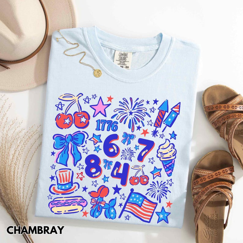 Puede incluir: Camiseta azul claro con un colorido dise&ntilde;o patri&oacute;tico que incluye el n&uacute;mero 1776, fuegos artificiales, cerezas, estrellas y una bandera estadounidense. La camiseta forma parte de una colecci&oacute;n de verano, mostrada con sandalias y un sombrero.