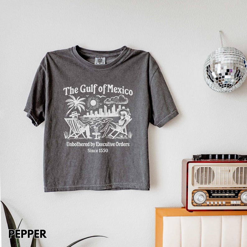 May include: A dark gray t-shirt with a white graphic print of a beach scene with two people sitting in lounge chairs. The text on the shirt reads "The Gulf of Mexico" and "Unbothered by Executive Orders Since 1550".
