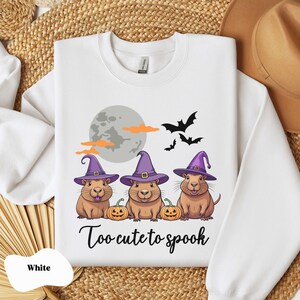 Op de afbeelding: Wit sweatshirt met een afbeelding van drie cartoon capybara's die toverhoeden dragen en pompoenen vasthouden. De tekst "Too cute to spook" is onder de capybara's gedrukt.